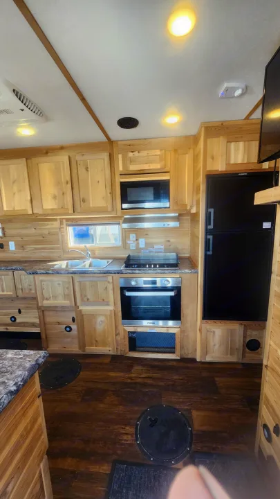 Inside an RV rental, a well-equipped kitchen with natural wood cabinets, modern appliances, and unique floor access hatches.. Ice Castle Fish Houses Toy Hauler 2022