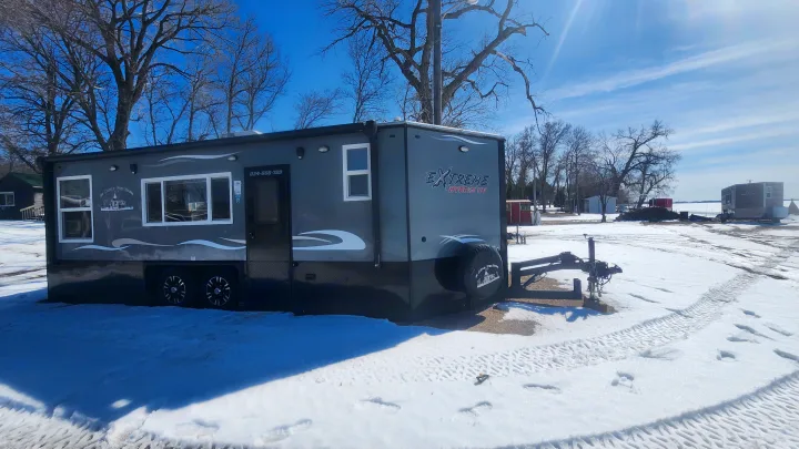 This 'Extreme Hybrid RV' fish house, available for rv rental, sits in a snowy winter landscape under a bright blue sky.. Ice Castle Fish Houses Toy Hauler 2022