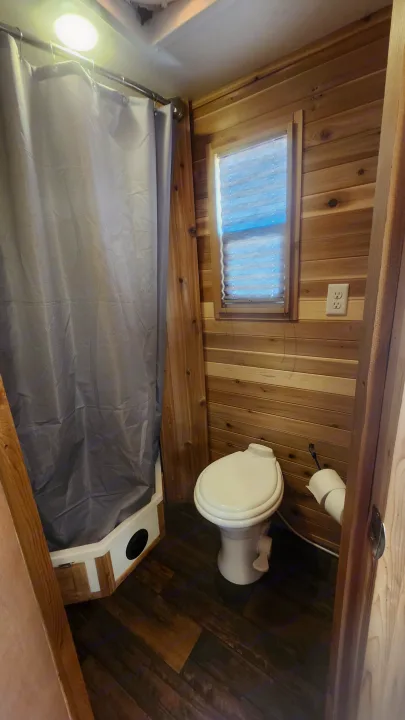This RV rental features a compact bathroom with warm wood paneling, a shower with a gray curtain, and a toilet.. Ice Castle Fish Houses Toy Hauler 2022