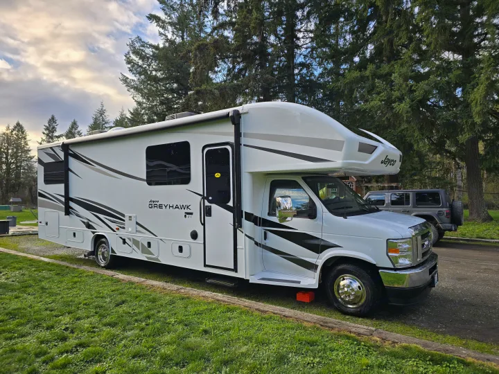 A white Jayco Greyhawk Class C RV rental, parked on grass at a peaceful campsite with tall trees in the background.. Jayco Greyhawk 31F Bunkhouse 2025