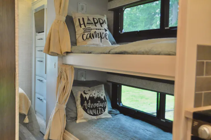 Bright RV rental interior featuring cozy bunk beds with "Happy Camper" and "Adventure Awaits" pillows, overlooking nature.. Jayco Greyhawk 31F Bunkhouse 2025