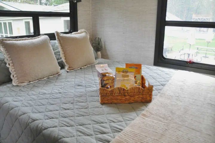 Cozy RV rental bed with light bedding, decorative pillows, and a welcome snack basket, offering a view of the outdoors.. Jayco Greyhawk 31F Bunkhouse 2025