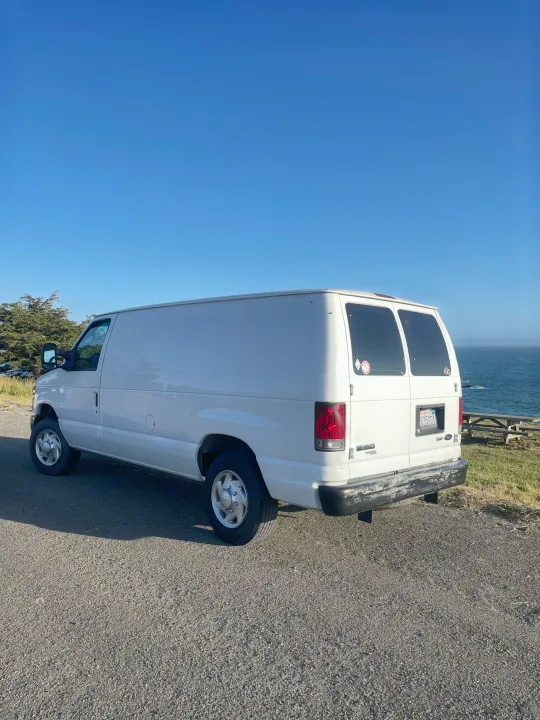 White Ford cargo van parked on a coastal road overlooking the Pacific Ocean. Perfect for an RV rental getaway.. Ford Econoline 150 2011