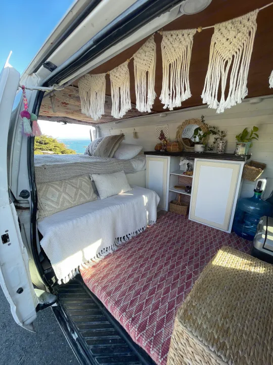 Cozy RV rental interior with boho decor, featuring a bed, seating, and an ocean view from the open back.. Ford Econoline 150 2011