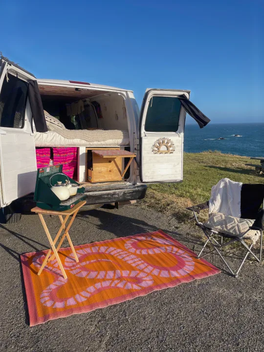 RV rental camper van parked by the ocean with an outdoor cooking setup, chair, and open doors showing the cozy interior.. Ford Econoline 150 2011