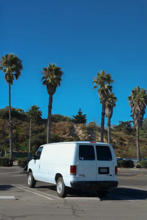 A white Ford E-150 van, a popular base for custom camper or RV rental, parked on a sunny day with tall palm trees and a clear sky.. Ford Econoline 150 2011