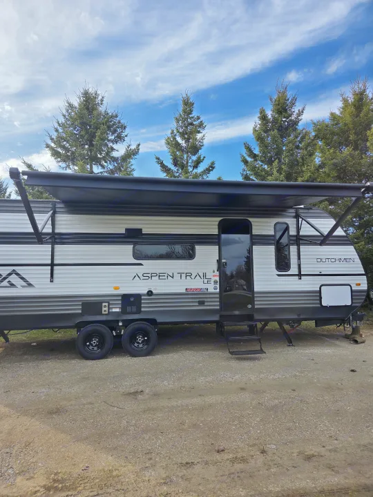 Spacious Aspen Trail LE travel trailer for your next RV rental adventure, parked on gravel with an extended awning under a blue sky.. Dutchmen Aspen Trail 2025