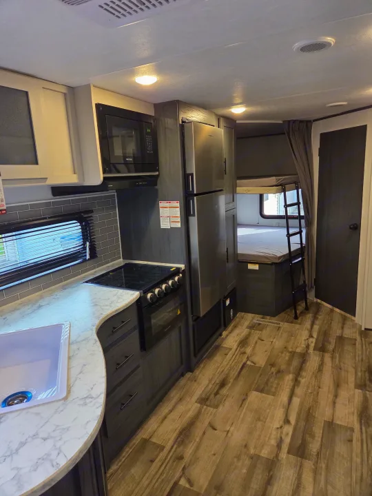 RV rental interior featuring a modern kitchen with stove, microwave, & fridge on the left, and bunk beds with a ladder on the right.. Dutchmen Aspen Trail 2025
