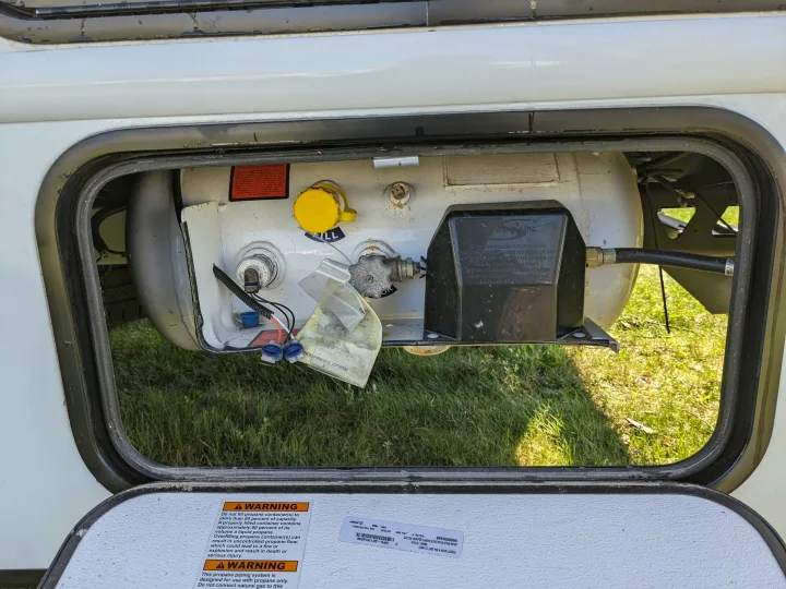 RV rental propane tank system with warning labels visible in an open utility compartment, showing the fill cap and valves.. East to West Alita 2025