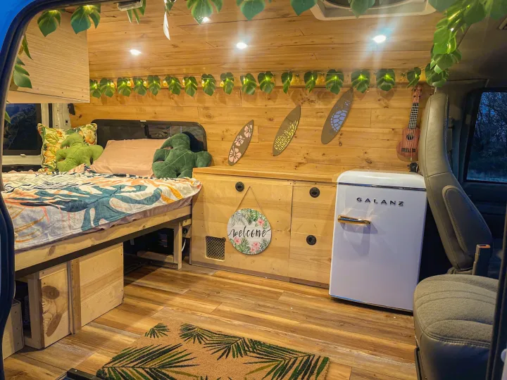 This RV rental interior features a cozy bed, mini-fridge, storage, and tropical decor with string lights & surfboards.. Chevrolet Express 1999
