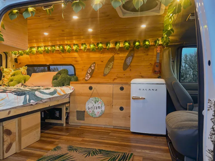 Cozy RV rental interior with wooden walls, bed, string lights, mini-fridge, and ukulele. Perfect for your next adventure.. Chevrolet Express 1999