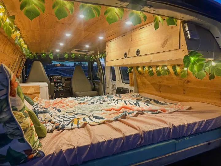 This cozy RV rental features a custom wood interior, complete with a comfortable bed, overhead cabinets, and tropical leaf string lights.. Chevrolet Express 1999