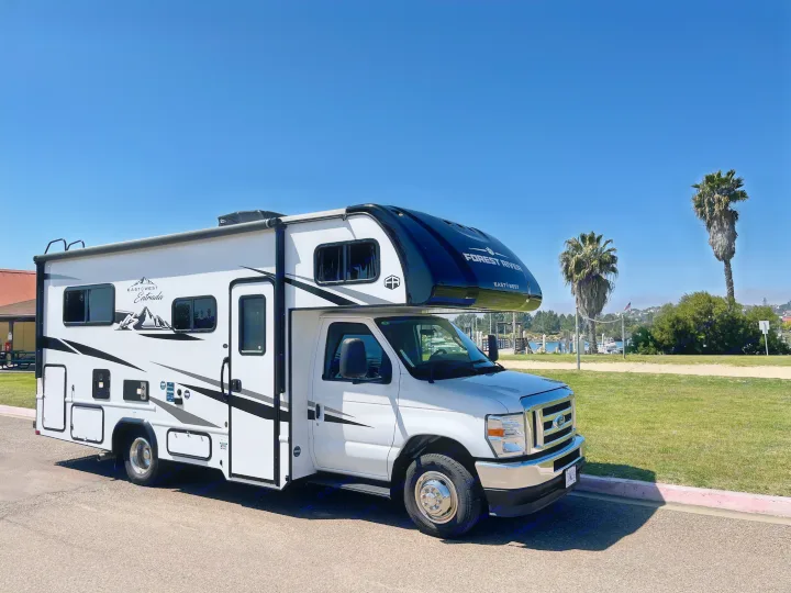 White Forest River Class C RV rental parked on pavement near a marina with palm trees under a sunny sky.. East to West Entrada 210NS 2025