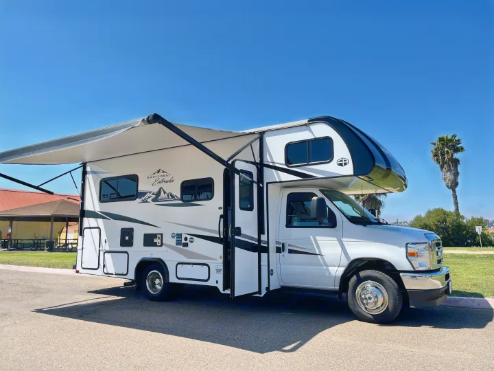 White Class C RV rental with an extended awning, parked on asphalt under a clear blue sky. Perfect for your next trip!. East to West Entrada 210NS 2025