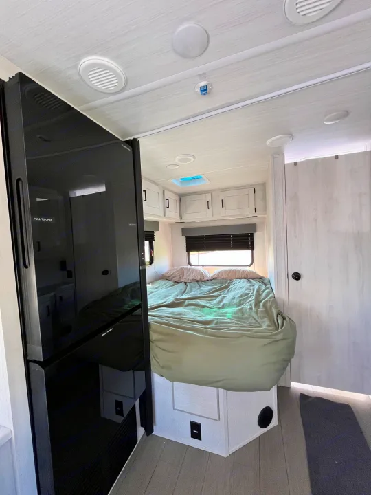 This RV rental's bright bedroom shows a comfortable bed, overhead cabinets, and a tall dark refrigerator unit.. East to West Entrada 210NS 2025