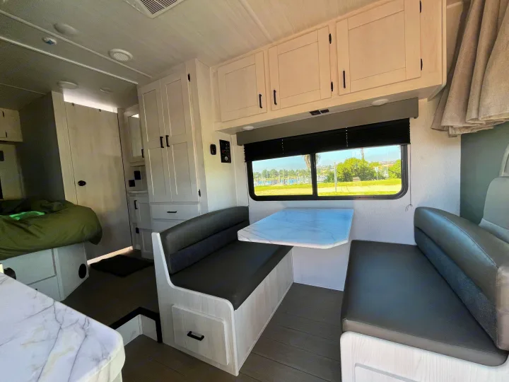 Spacious RV rental interior featuring a bright dining booth, overhead cabinets, and a glimpse of the bedroom. Enjoy the scenic window view.. East to West Entrada 210NS 2025