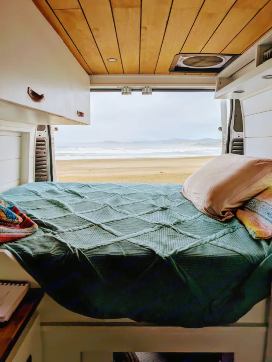 View from an RV rental's bed looking out to a sandy beach and ocean waves. Cozy van interior, wooden ceiling, and coastal scenery.. Dodge Ram 1500 2014