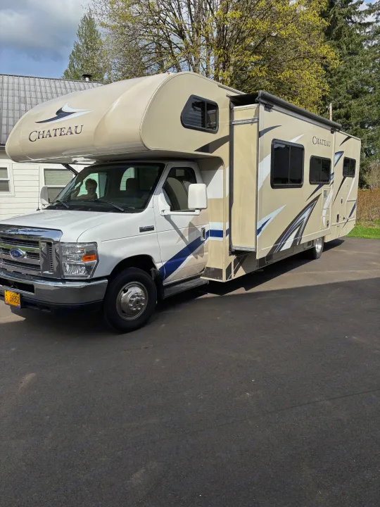 A white and beige Chateau RV rental is parked on an asphalt driveway, with a child visible in the driver's seat.. Thor Chateau 2019