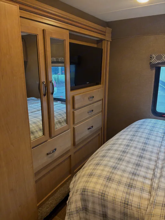 Cozy RV rental bedroom featuring built-in cabinetry, TV, and mirrored closet, with comfy plaid bedding.. Thor Chateau 2019