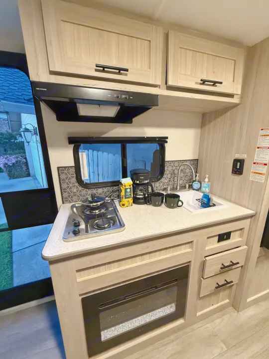 A clean RV rental kitchen interior, with a stove, sink, coffee maker, light wood cabinets, and an electric fireplace.. Heartland Trail Runner 2025