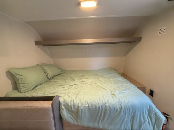 Compact rv rental bedroom with a bed, light green bedding, overhead shelf, and an LCD TV mounting label.. Heartland Trail Runner 2025