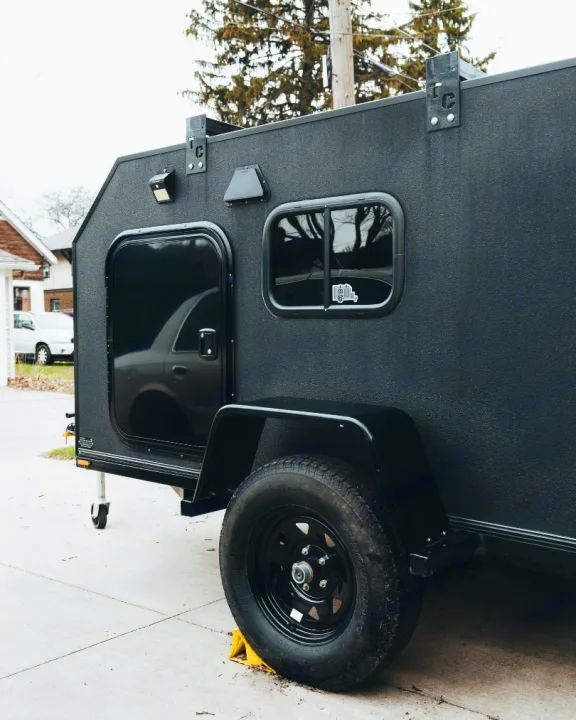 Close-up of a compact, black textured overland trailer, ideal for RV rental adventures. Features a door, window, and rugged wheel.. Other Other 2022