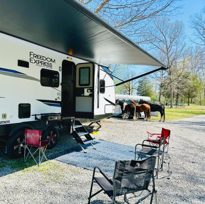 RV rental parked at a serene campsite with awning, outdoor rug, chairs, and horses in background. Ideal for nature lovers.. Forest River Coachmen Freedom Express 2025