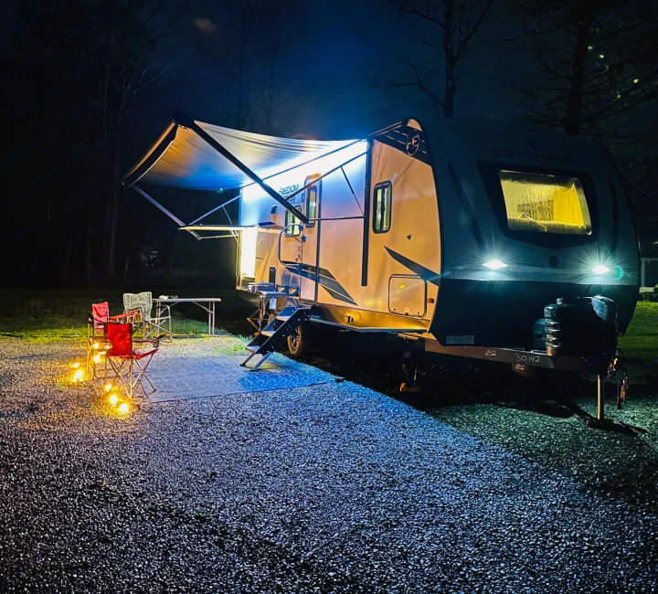 Cozy RV rental setup at a night campsite. The illuminated travel trailer has an extended awning, chairs, and warm ground lights.. Forest River Coachmen Freedom Express 2025