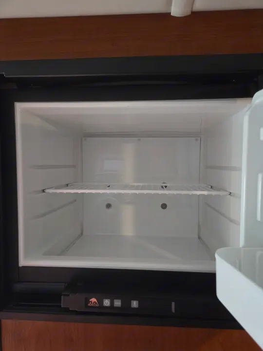 A clean, empty RV rental refrigerator is shown, with its door open, revealing a wire shelf and a Norcold control panel below.. Winnebago Sunstar 2019