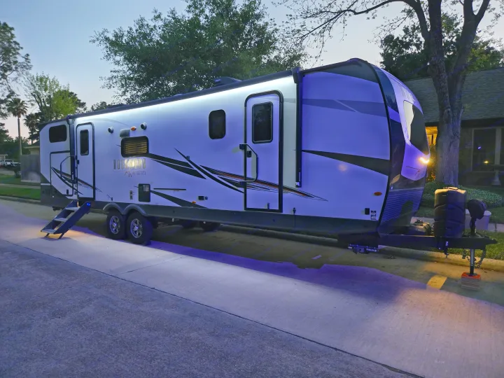A modern white and black travel trailer, brightly lit with blue/white LEDs at dusk, parked curbside. Explore an rv rental adventure.. Forest River Rockwood Ultra Lite 2022
