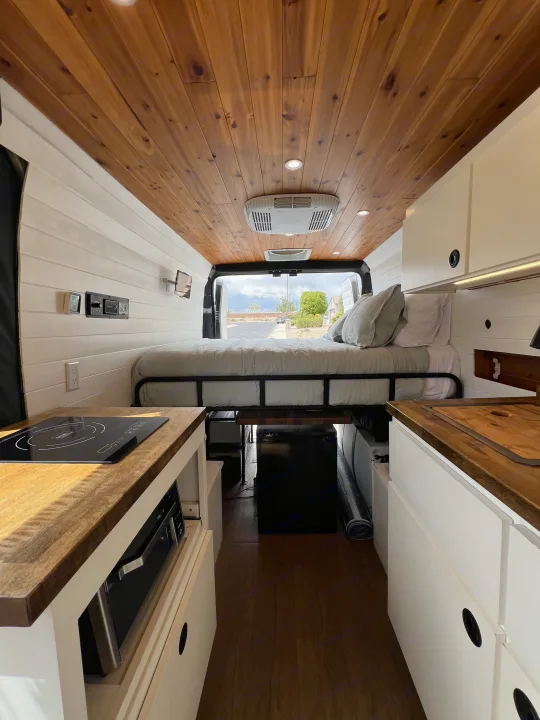 Inside a stylish RV rental van. Shows a compact kitchen, elevated bed, wood ceiling, and white paneling.. Dodge Ram Promaster 2500 2022