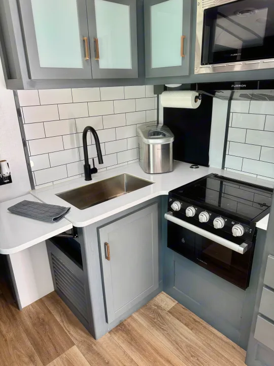 Modern RV rental kitchen with gray cabinets, subway tile, sink, Furrion oven, microwave, and Frigidaire ice maker.. Keystone RV Bullet Premier Ultra Lite 2022