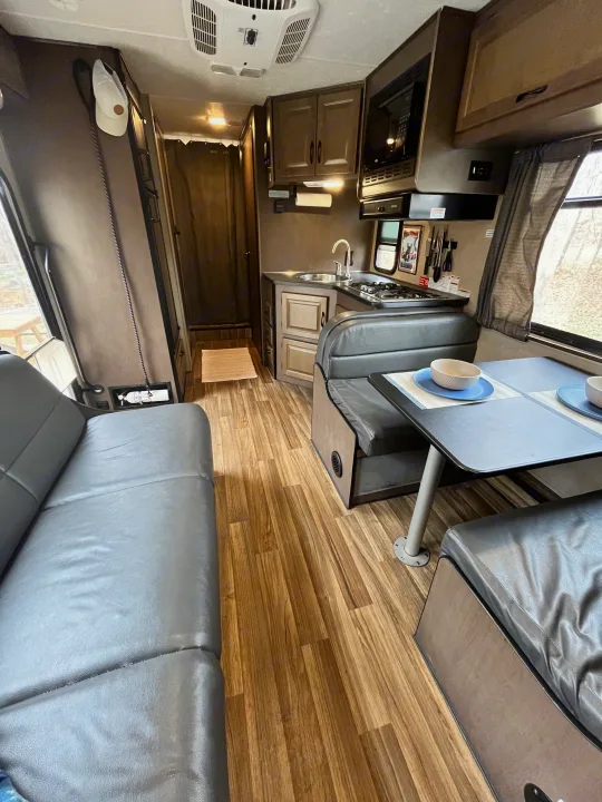 View inside an rv rental, showing a gray sofa, dinette with two place settings, and a compact kitchen with wood floors.. Thor Majestic 2019