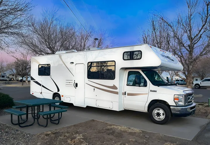 A white RV rental, a Class C motorhome, is parked at a scenic campsite with a green picnic table, under bare trees and a blue sky.. Thor Majestic 2019