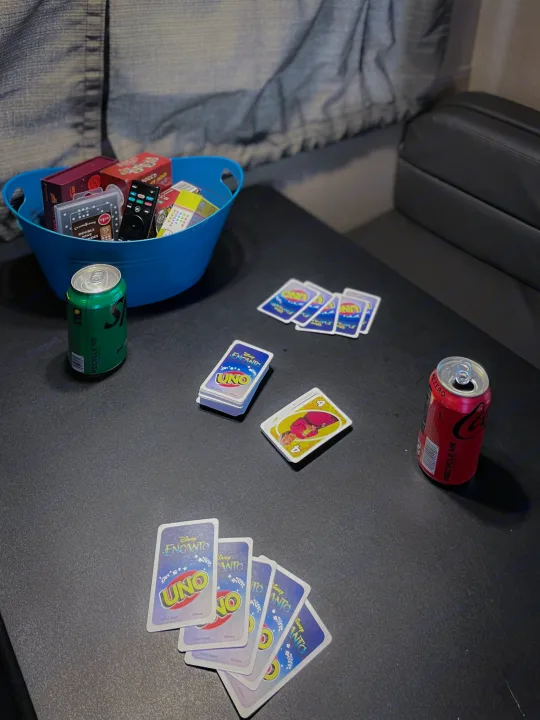 RV rental entertainment: A blue basket of games & remotes, Disney Encanto UNO cards, and two soda cans on a table.. Thor Majestic 2019