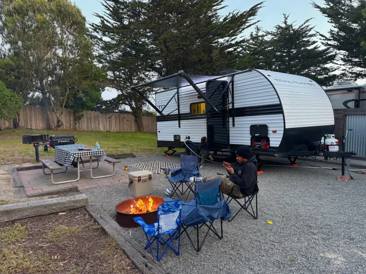 Cozy campsite scene with an RV rental, a man relaxing by the fire pit, and a child near the trailer.. Heartland Trail Runner 2025