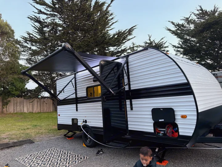 Our spacious RV rental set up at a peaceful campsite, with an extended awning and a child playing on the mat.. Heartland Trail Runner 2025