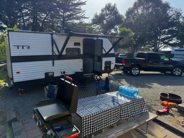 Enjoying an RV rental setup! A white travel trailer, black pickup, grill, table, and chairs at a sunny campsite.. Heartland Trail Runner 2025