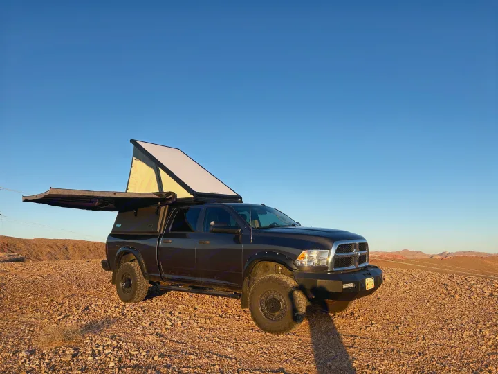 A black overlanding truck with a deployed pop-top tent and side awning, parked on rocky desert ground. An adventurous alternative to an RV rental.. Dodge Dodge 1 Ton Pu 2018