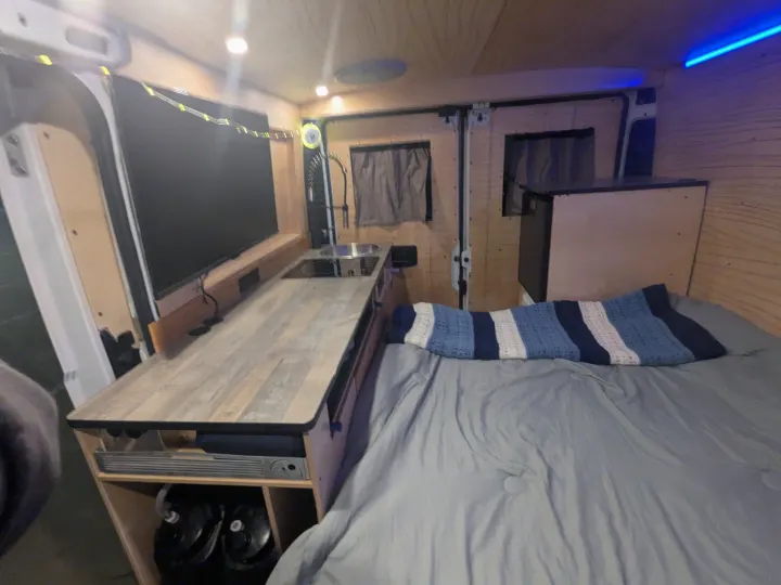 Cozy RV rental interior with wood paneling, kitchenette, mounted TV, and a snug bed with blue accent lighting.. RAM Promaster 2023