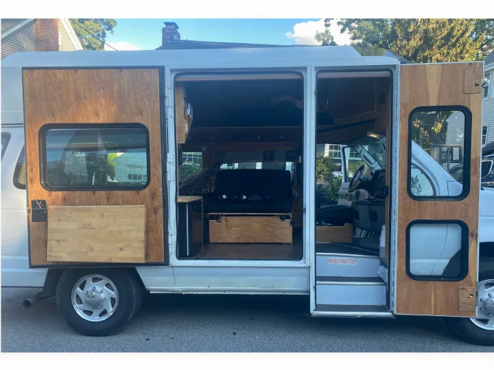 Detailed view of a white camper van's custom wooden interior, accessible through open side doors. Explore RV rental options!. Ford Econoline 2011