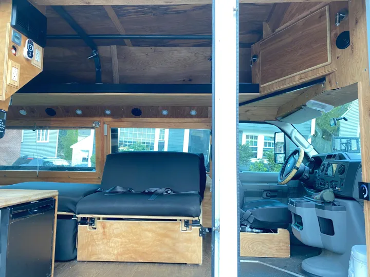Custom-built RV rental camper van interior featuring wooden cabinetry, convertible seating, a compact fridge, and overhead bed platform.. Ford Econoline 2011