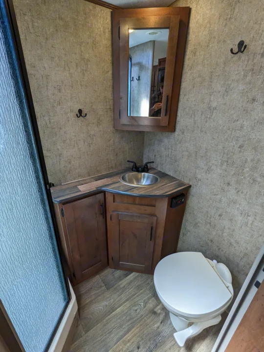 Compact RV rental bathroom with corner sink, toilet, and shower. Features rustic wood cabinets and textured walls.. Outdoors RV Timber Ridge 2022