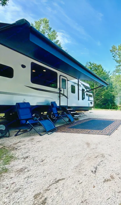 Relaxing at an RV rental campsite. White RV with extended awning, blue chairs, and an outdoor mat on gravel under a blue sky.. Forest River Coachmen Northern Spirit Ultra Lite 2023