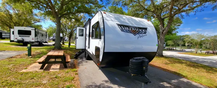 A white Wildwood Platinum RV rental is parked at a scenic campground site with a picnic table, ideal for outdoor adventures.. Forest River Wildwood Platinum 2024