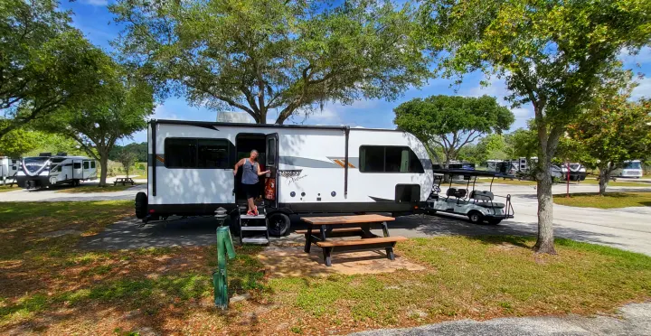 Sunny RV rental campsite: White trailer with a person, picnic table, and golf cart. Other RVs dot the background.. Forest River Wildwood Platinum 2024