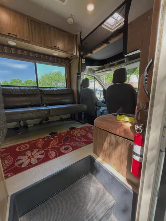 RV rental interior view: spacious with dark leather seating, wood cabinets, and a patterned rug. Cab area & fire extinguisher seen.. Forest River Coachmen Freelander Micro 2018