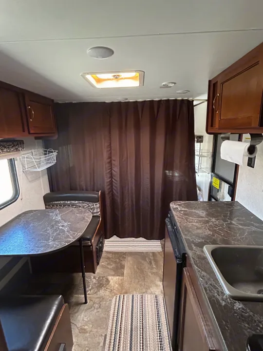 This RV rental's interior features a compact kitchen counter with a sink and a dining booth with a table, separated by brown curtains.. Jayco Jay Flight SLX Baja Edition 2017