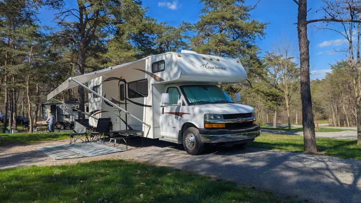 A white RV rental is perfectly set up at a sunny campsite on a gravel pad, complete with an extended awning and outdoor seating.. Forest River Coachmen Freelander 2013