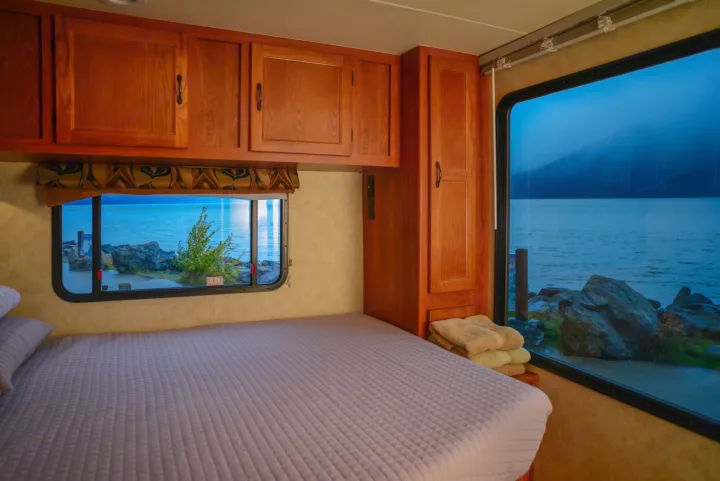 Inside an RV rental bedroom overlooking a tranquil lake and rocky shore at dusk. Enjoy peaceful views from your bed.. Forest River Coachmen Freelander 2014